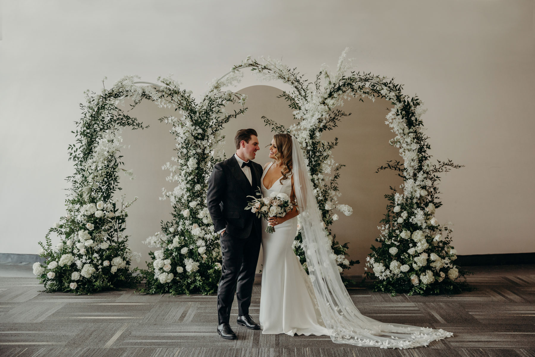 A bride and groom stand in front of floral arches.