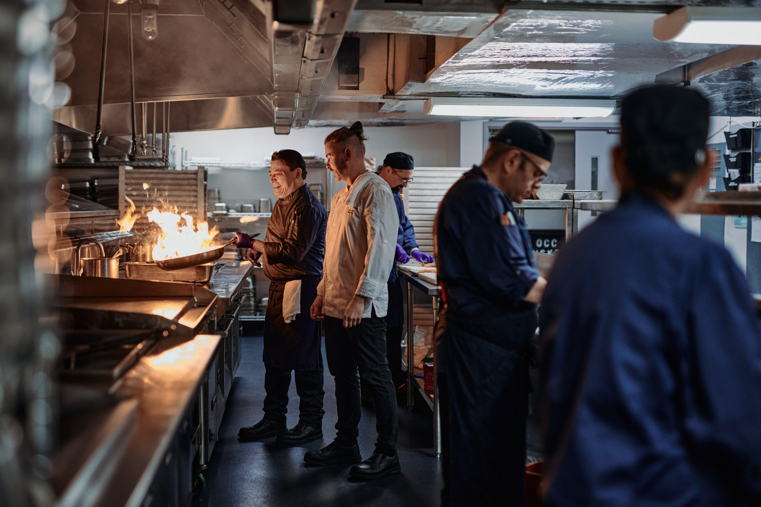 people in large industrial kitchen around a stove with a flame